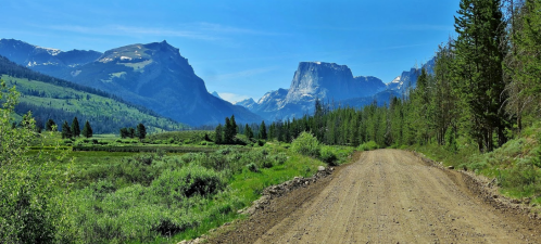 A dirt road winds through a lush green landscape, with mountains and blue skies in the background.