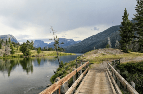 A wooden bridge leads to a serene lake surrounded by mountains and trees under a cloudy sky.