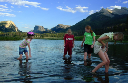 Four children play in a shallow river, surrounded by mountains and a clear blue sky.
