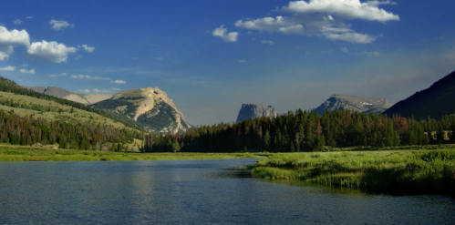 A serene landscape featuring a calm river, lush greenery, and mountains under a blue sky with scattered clouds.