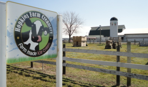 Sign for Hopkins Farm Creamery with a cow logo, set in a rural landscape featuring a barn and fenced area.