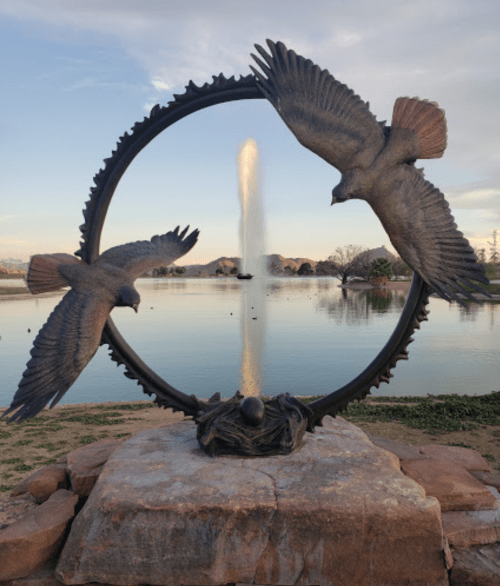 A bronze sculpture of two birds flying around a circular frame, with a fountain and lake in the background.