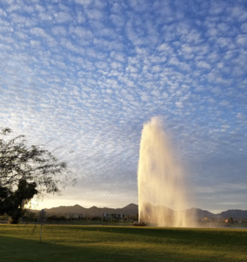 A tall water fountain sprays into a blue sky with scattered clouds, set against a backdrop of mountains and green grass.