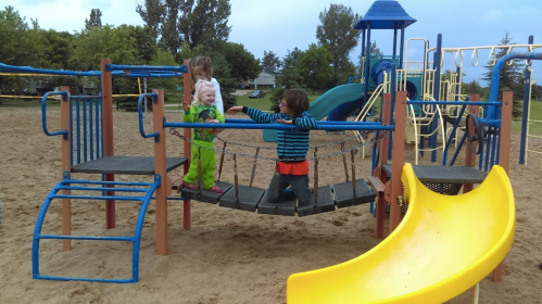 Three children play on a playground bridge, with a slide and climbing structures in a sandy area.