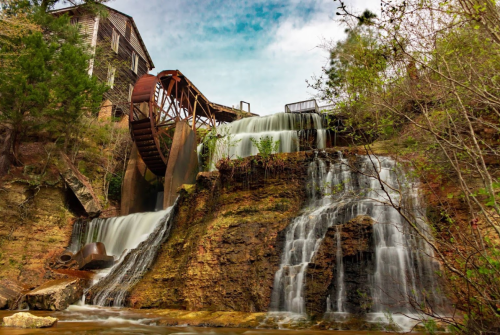 A scenic view of a watermill with a large wheel, cascading waterfalls, and lush greenery under a blue sky.