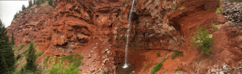 A waterfall cascades down red rock formations, surrounded by greenery in a scenic landscape.