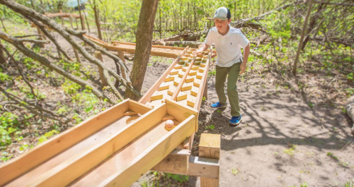A child plays with a wooden marble run in a forested area, smiling as he interacts with the structure.