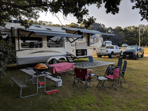 A spacious RV parked on grass, with picnic tables, chairs, and a grill set up for outdoor relaxation.