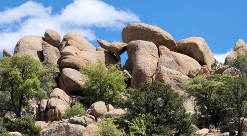 A cluster of large, weathered boulders surrounded by green shrubs under a bright blue sky with scattered clouds.