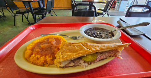 A plate with a Cuban sandwich, yellow rice, and black beans on a red tray in a casual dining setting.