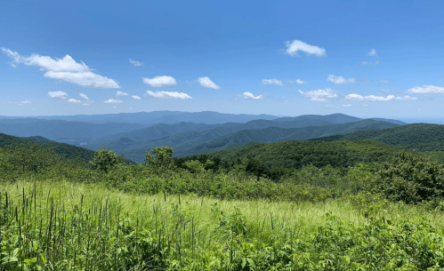 A panoramic view of rolling green mountains under a clear blue sky with scattered clouds.