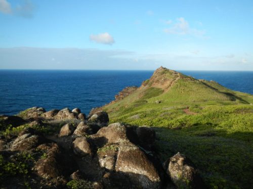 A rocky hillside leads to a peak overlooking a calm blue ocean under a clear sky.