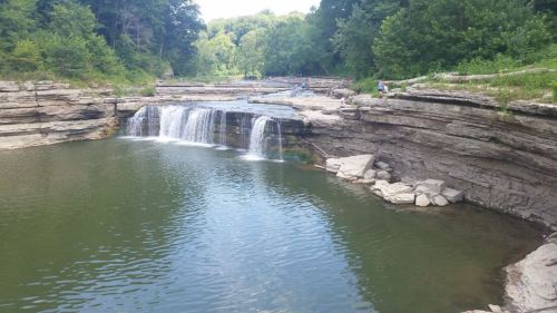 A serene waterfall cascades into a calm pool, surrounded by lush greenery and rocky cliffs.