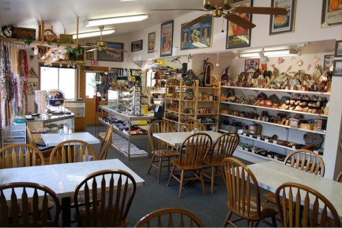 Cozy café interior with wooden tables, chairs, and shelves displaying various items and decorations. Bright and inviting atmosphere.