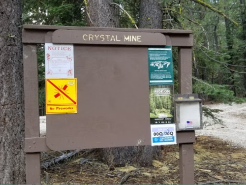 Signboard at Crystal Mine with notices, including no fireworks and various informational flyers, surrounded by trees.