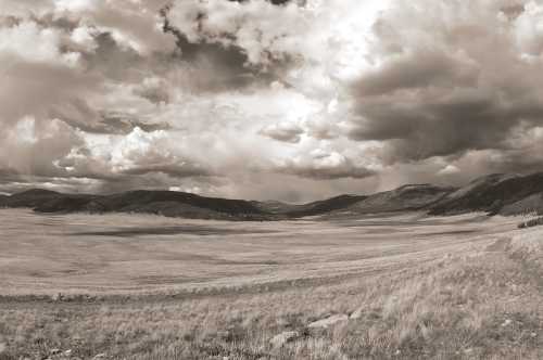 A panoramic view of a vast, open landscape with rolling hills and dramatic, cloudy skies in black and white.