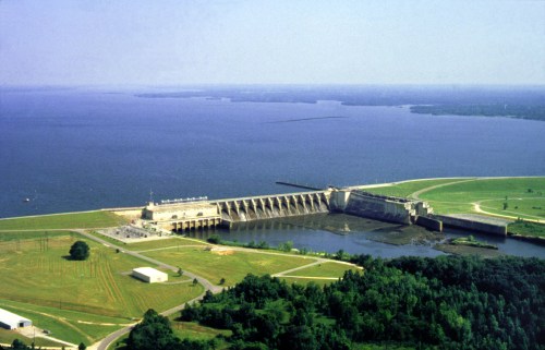 Aerial view of a large dam with water reservoir, surrounded by green fields and trees under a clear blue sky.