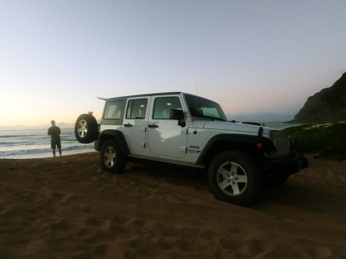 A white SUV parked on sandy beach with a person standing by the water, mountains in the background at sunset.