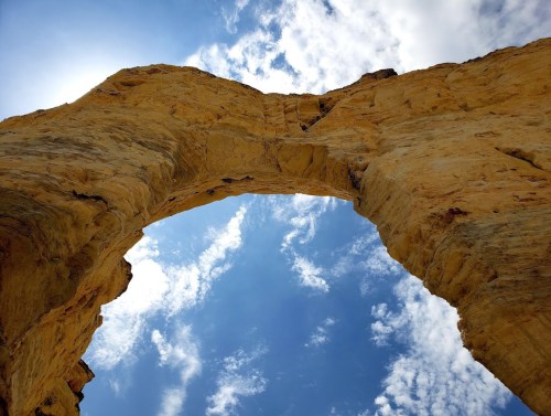 View from below a large rock arch, framed by a bright blue sky with scattered clouds.