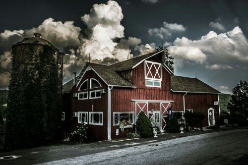 A red barn with a silo under a dramatic sky filled with clouds, surrounded by greenery.