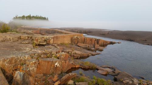 A rocky shoreline with a calm river, fog rolling in over a distant island surrounded by trees.