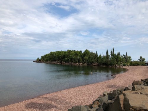 A serene lakeside view with a sandy beach, rocky shore, and lush green island under a cloudy sky.