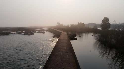 A foggy landscape featuring a narrow path over water, surrounded by trees and distant hills.