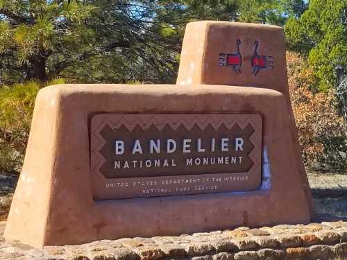 Sign for Bandelier National Monument, featuring adobe-style design and colorful symbols, surrounded by trees.