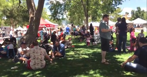 A lively outdoor event with people sitting on grass, enjoying food and socializing under trees and tents.