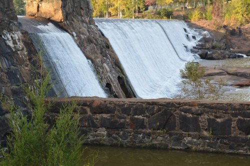 A waterfall cascading over a stone dam, surrounded by greenery and rocky terrain.