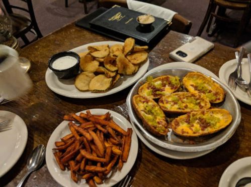 A table with three plates of food: sweet potato fries, potato skins topped with cheese, and crispy potato chips with dip.