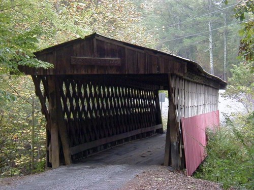 A rustic wooden covered bridge surrounded by trees, with a gravel path leading through it.
