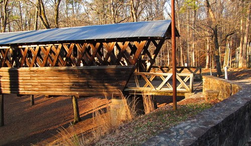 A wooden covered bridge with a metal roof, surrounded by trees in a wooded area during late afternoon light.
