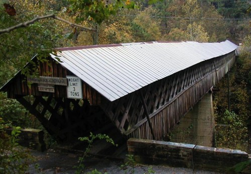 A wooden covered bridge spans a small river, surrounded by trees with autumn foliage. A weight limit sign is visible.