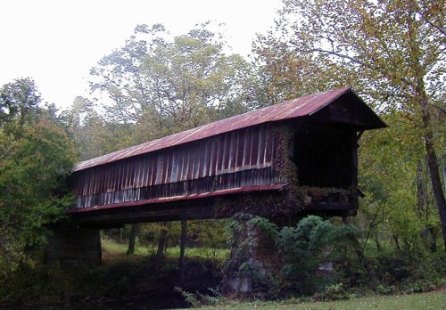 A rustic covered bridge with a red roof, surrounded by trees and greenery, spanning a small creek.