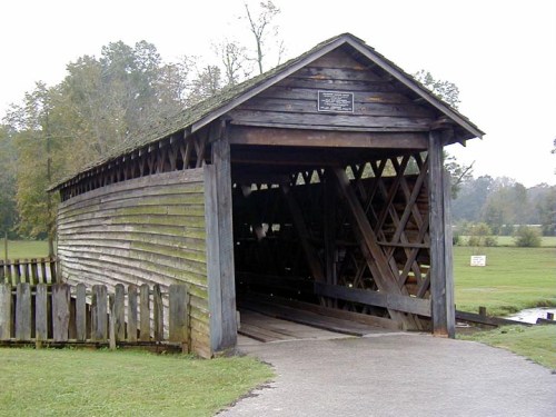A rustic wooden covered bridge with a sloped roof, surrounded by greenery and a gravel path leading to its entrance.