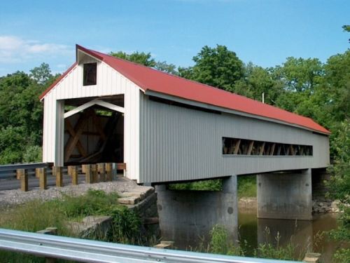 A white covered bridge with a red roof spans over a river, surrounded by greenery and a gravel road.