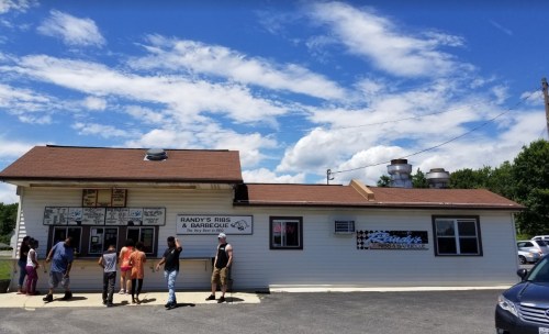 A barbecue restaurant with a brown roof and people lined up outside under a blue sky with clouds.