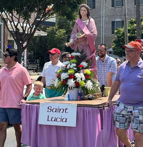 A group of people carrying a statue of Saint Lucia on a decorated float during a parade, surrounded by flowers.