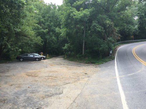 A gravel parking area next to a winding road, surrounded by dense greenery and trees.