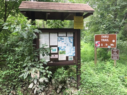 Information board at a trailhead surrounded by greenery, with signs for a thru trail and an adopt-a-trail program.