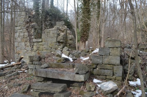 Ruins of a stone structure surrounded by trees and scattered snow, with remnants of walls and steps visible.
