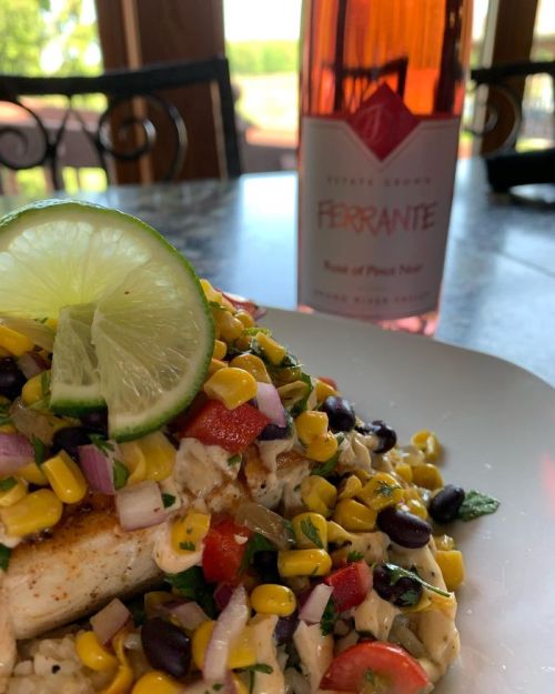 A plate of grilled fish topped with corn, tomatoes, and onions, alongside a bottle of rosé wine and a lime wedge.