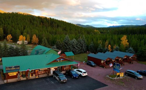 Aerial view of a rustic general store with cabins nearby, surrounded by trees and mountains under a cloudy sky.