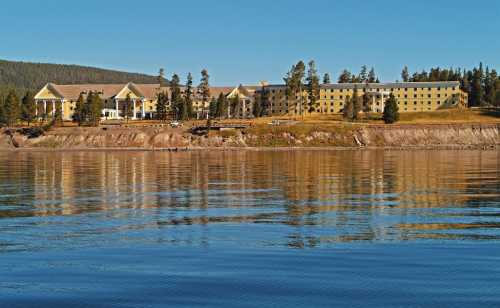 A lakeside hotel surrounded by trees, reflecting in calm water under a clear blue sky.