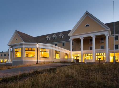 A large, yellow hotel building with illuminated windows, set against a twilight sky and surrounded by grass.