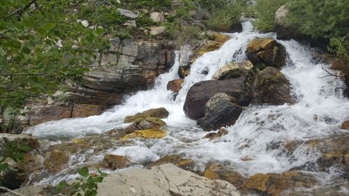 A cascading waterfall flows over rocks, surrounded by lush greenery and natural scenery.