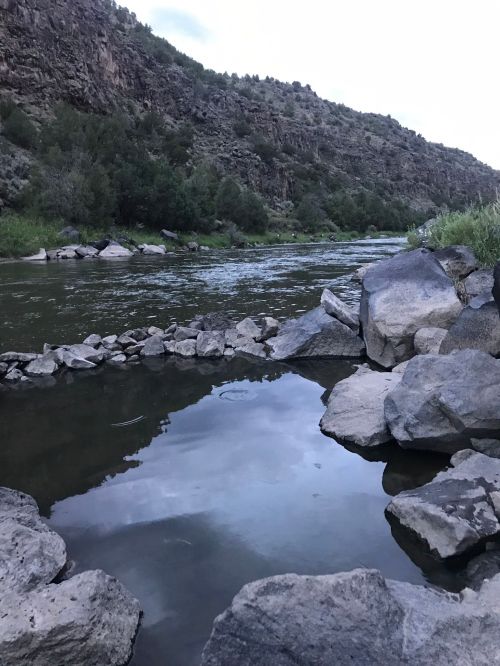 A serene river flows between rocky banks, reflecting the sky and surrounding greenery in a peaceful landscape.