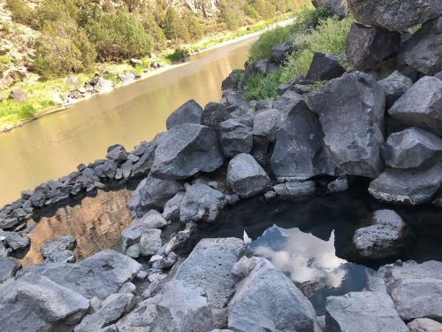 A natural hot spring surrounded by large rocks, with a river and greenery in the background.