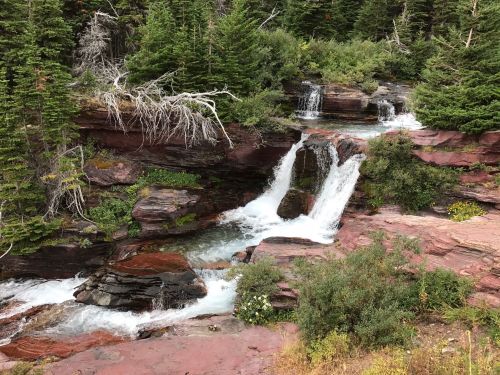A serene waterfall cascades over red rock formations, surrounded by lush green trees and foliage.